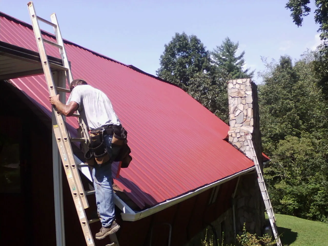 Roofer on a ladder beside a red metal roof and stone chimney — Soffit & Fascia Repair by Roofing Brothers in Dix